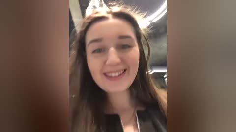 Media: A video of a young Caucasian woman with long brown hair and fair skin, smiling. She wears a black leather jacket. The background is a dimly lit, modern room with a circular ceiling light.