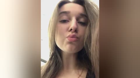 Media: Video of a young Caucasian woman with straight, shoulder-length blonde hair, making a pouty face. She has a fair complexion, minimal makeup, and wears a black top. Background is a beige wall, indoor setting.