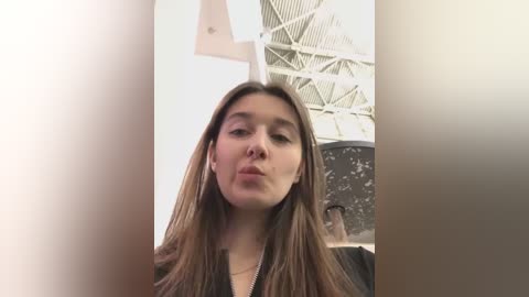 Media: Video of a young woman with long brown hair, fair skin, and a straight face, taken from a low angle, showing the ceiling with white beams and a metal sculpture in the background.