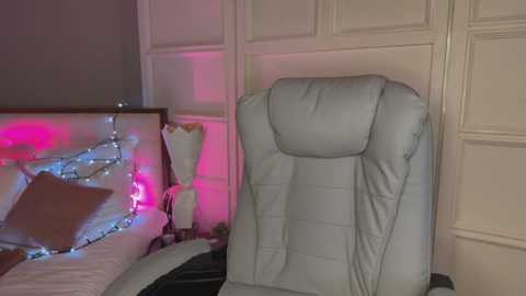 Media: Video of a cozy bedroom with a white leather recliner chair, a lit string of blue and pink lights, and a wooden headboard with a plush pillow and a beige blanket.