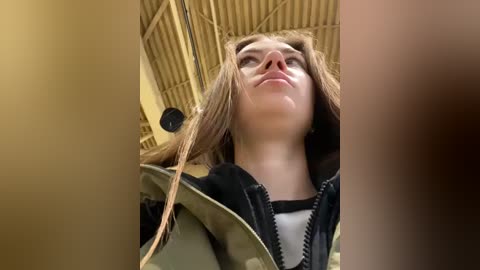 Media: Video of a young Caucasian woman with long brown hair, wearing a black jacket, looking upward, in a dimly lit, corrugated metal-roofed room.