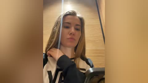 Media: Video of a young Caucasian woman with long blonde hair, wearing a black and white striped sweater, reflected in a mirror, with wooden walls in the background.