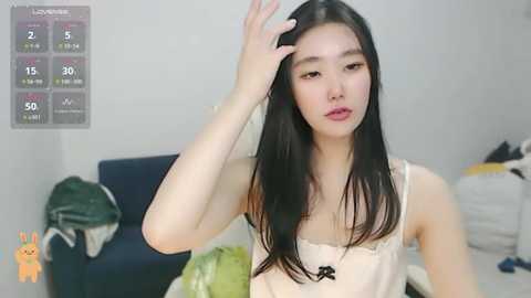 Media: Video of a young Asian woman with long black hair, wearing a white spaghetti-strap top, adjusting her hair in a messy room with a calendar and clothes scattered on the floor.