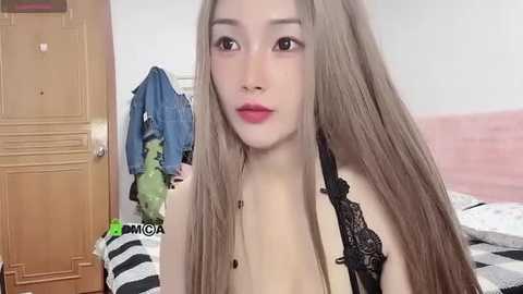 Media: A video of an Asian woman with long, straight, light brown hair, fair skin, and red lipstick, wearing a black lace bra, in a bedroom with a wooden door, green and blue clothes, and a pink wall.