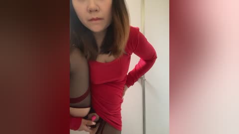 Media: Video of a fair-skinned, petite Asian woman with shoulder-length brown hair, wearing a tight red top, standing in a room with white walls and a door. She's holding a pink vibrator between her legs.