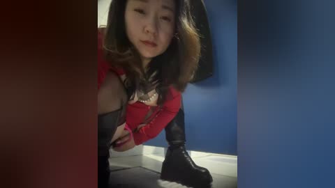 Media: Video of a young Asian woman with long black hair, wearing a red top, black leather jacket, and black boots, bending down in a dimly lit, blue-walled room.