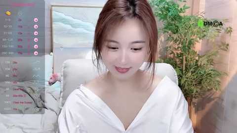 Media: A video of an Asian woman with light skin, wearing a white robe, sitting indoors. Background features a potted plant and a bed with white bedding.