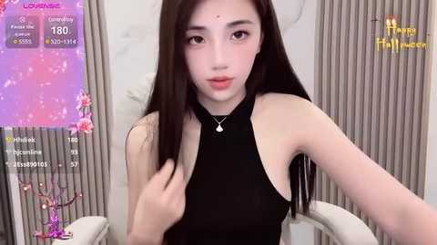 Media: Video of a young Asian woman with long black hair, fair skin, and a slim physique, wearing a black halter top, sitting in a modern room with a pink floral screen and a live stream overlay.