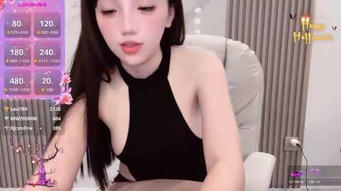 Media: Video of an Asian woman with long black hair in a black halter top, sitting in a modern office chair. The background features grey curtains and a digital display showing viewer stats.