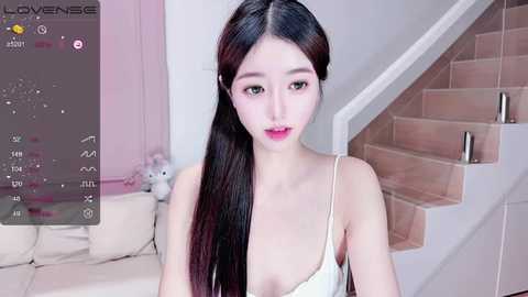 Media: Video of an East Asian woman with long black hair, fair skin, and red lipstick, wearing a white spaghetti-strap top, standing in a modern, minimalist room with a beige sofa, plush toys, and wooden stairs.