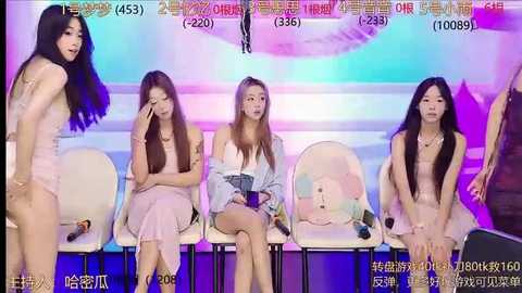 Media: Video of four young women with long hair, sitting on white chairs in a colorful studio. They wear casual outfits, one woman has a pink shirt. Background shows a light-up sign.