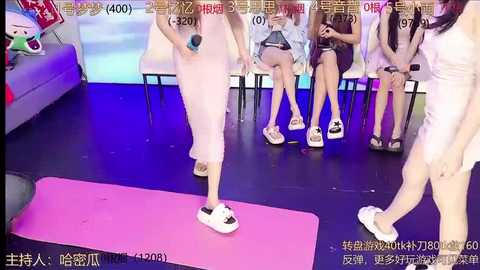 Media: Video of four Asian women in white sportswear, seated on chairs, with a woman in white slippers center stage. Background includes a pink yoga mat, colorful banners, and a gray sofa.