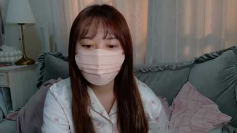 Media: Video of an Asian woman with long brown hair and pale skin, wearing a pink surgical mask, white pajamas, and sitting on a gray couch with pink pillows, in a cozy, dimly lit room.