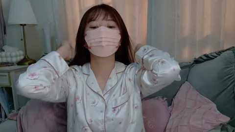 Media: Video of a young Asian woman with long brown hair, wearing a pink surgical mask and white pajamas, adjusting her hair in a cozy living room with pastel cushions and a lamp.