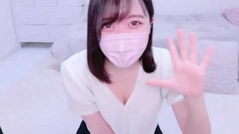 Media: Video of a young East Asian woman with light skin and shoulder-length dark hair, wearing a white V-neck blouse, pink mask, and waving from a white room with a fluffy carpet.