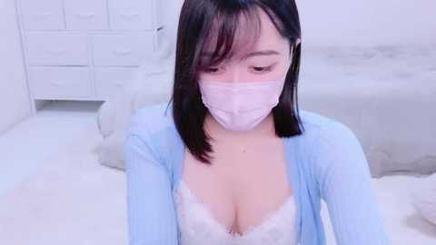 Media: A video of an Asian woman with straight black hair, wearing a light blue cardigan, white lace bra, and pink face mask, sitting on a white bed, in a minimalistic, white room.