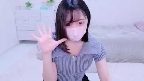 Media: A video of an East Asian woman with straight black hair, wearing a face mask, blue and white striped top, and a white hand raised, standing in a bright, minimalistic bedroom with white walls and a bed.