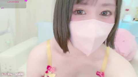 Media: Video of a young Asian woman with short brown hair, wearing a pink face mask, yellow spaghetti-strap top adorned with pink and red flowers. She has a soft, light skin tone and is indoors, likely a bedroom setting.