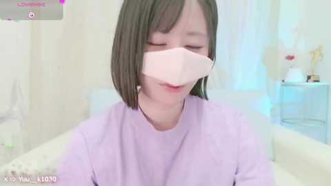 Media: Video of an East Asian woman with short brown hair wearing a light lavender top and white face mask, smiling gently, in a bright, minimalistic room with white walls and a window.