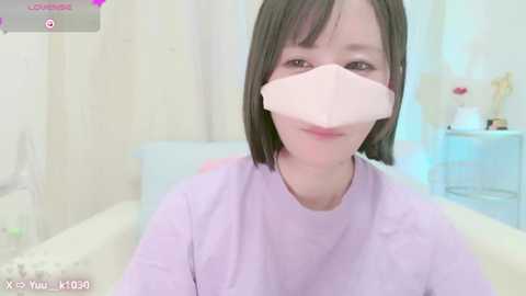 Media: A video of a young Asian woman with straight black hair and light skin, wearing a light purple shirt and a pink surgical mask, in a hospital room with white walls and a bed.