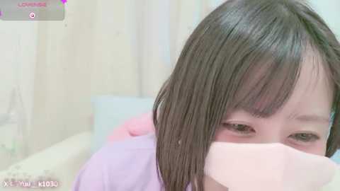 Media: Video of an Asian woman with straight, shoulder-length brown hair, partially obscured by a pink surgical mask. She wears a light purple shirt. Background features a blurred, pale curtain.