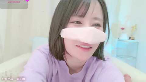 Media: A video of a young Asian woman with short black hair, wearing a lavender shirt and a pink surgical mask, smiling in a bright, modern bathroom with white walls and a mirror.