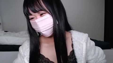 Media: Video of an East Asian woman with long black hair, wearing a pink surgical mask, black lace bra, and white fuzzy robe, indoors with a white wall and bed in the background.