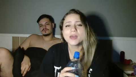 Media: Video of a woman drinking water in a black shirt and a man asleep in a dark towel, both sitting on a bed with beige walls and a red object on a nightstand.