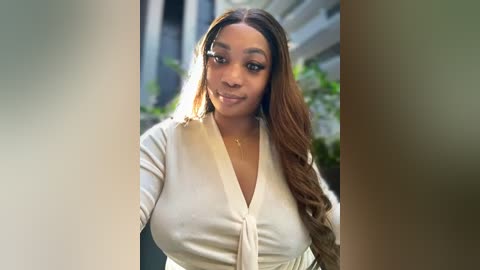 Media: Video of a Black woman with long, wavy brown hair, wearing a white V-neck cardigan, standing outside near a building with greenery in the background.