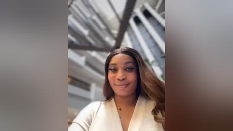 Media: Video of a young Black woman with a medium complexion, long wavy hair, and a warm smile, wearing a white cardigan, standing in a modern, industrial-looking room with white walls and exposed beams.