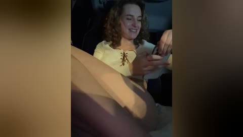 Media: A video of a smiling, curly-haired woman in a white top, sitting in a dimly lit car, holding a phone. The background is blurred.