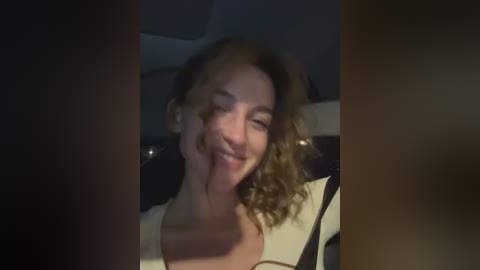 Media: Video of a young woman with curly hair, smiling and sticking her tongue out, wearing a white top, in a dimly lit car interior.