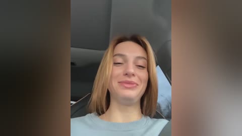 Media: Video of a young woman with shoulder-length, light brown hair, fair skin, and a slight smile, seated in a car with gray upholstery.