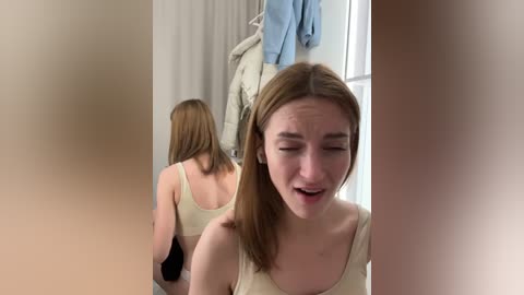 Media: Video of a young woman with light skin and straight, shoulder-length brown hair, wearing a yellow tank top, standing in a bright room with beige walls and a window. She appears startled, eyes closed, mouth open.