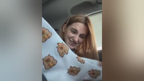 Media: Video of a young woman with long, straight, light brown hair, smiling inside a car with a white pillowcase featuring cartoon hamburger designs.