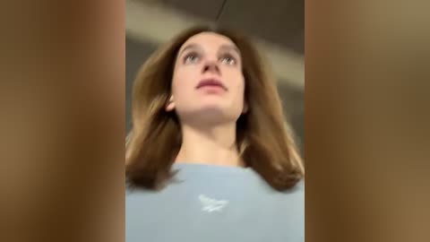 Media: Video of a young woman with shoulder-length brown hair, wearing a light blue shirt, looking up with a thoughtful expression. The background is blurred, suggesting an indoor setting.