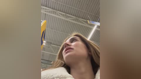 Media: Video of a young Caucasian woman with long brown hair, wearing a white furry jacket, looking upward in an industrial setting with a corrugated metal roof and a yellow plane propeller in the background.