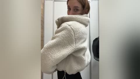 Media: A video of a young woman with light skin and straight, brown hair, partially hidden behind a white, fuzzy, hooded jacket, standing in a narrow hallway with beige walls and a white door.