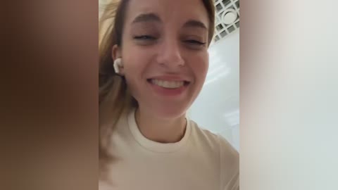 Media: A close-up video of a young woman with light skin and brown hair, smiling, wearing a white t-shirt. The background shows a white ceiling and a blurry window.