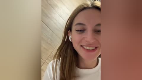 Media: Video of a young woman with straight brown hair, wearing white headphones, smiling in a narrow, wooden-paneled hallway.