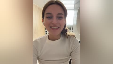 Media: Video of a young Caucasian woman with light skin and brown hair tied back. She's smiling, wearing a white long-sleeve shirt. Background shows a white wall and part of a room with a window and furniture.