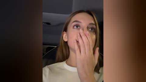 Media: Video of a young woman with light skin and straight brown hair, wearing a white shirt, covering her mouth with her left hand, looking shocked in a dimly lit car interior.