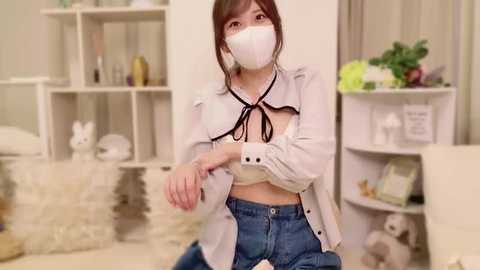 Media: Video of an Asian woman with light skin, wearing a white mask, white blouse, and blue jeans, seated in a modern, minimalist living room with white shelves and plants.