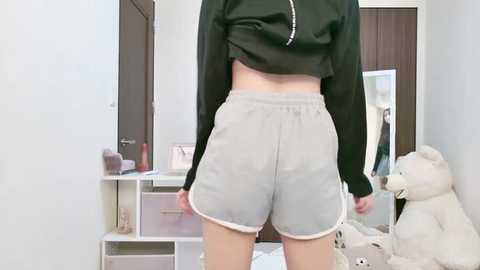 Media: A video of a woman standing in a well-lit, tidy bedroom. She wears a black crop top and light gray shorts, showcasing her midriff and slim legs. Background includes white shelves, a stuffed teddy bear, and closed brown doors.