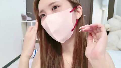 Media: A video of an Asian woman with long brown hair, wearing a pink surgical mask, adjusting the ear loops in a bright, modern bathroom.