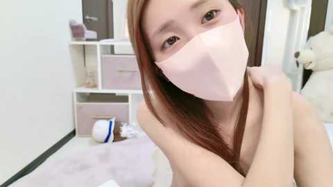 Media: Video of an Asian woman with long brown hair, wearing a pink face mask, sitting in a pastel-colored room with white shelves and a teddy bear.