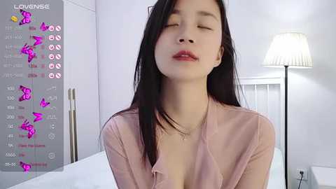 Media: Video of a young Asian woman with long black hair, wearing a pink blouse, sitting on a bed in a minimalist bedroom with a white lamp.