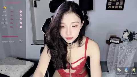 Media: Video of an Asian woman with long, wavy black hair, wearing a red lace bra and black choker, sitting in a gaming chair in a room with a white wall and framed pictures.