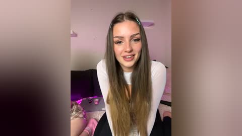 Media: Video of a smiling young woman with long, straight brown hair, wearing a white top and black pants, seated on a bed with pink sheets.