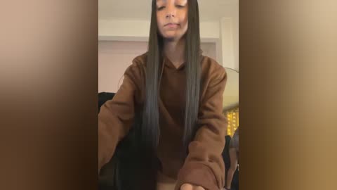 Media: Video of a woman with long black hair, wearing a brown hoodie, sitting indoors, blurry background.